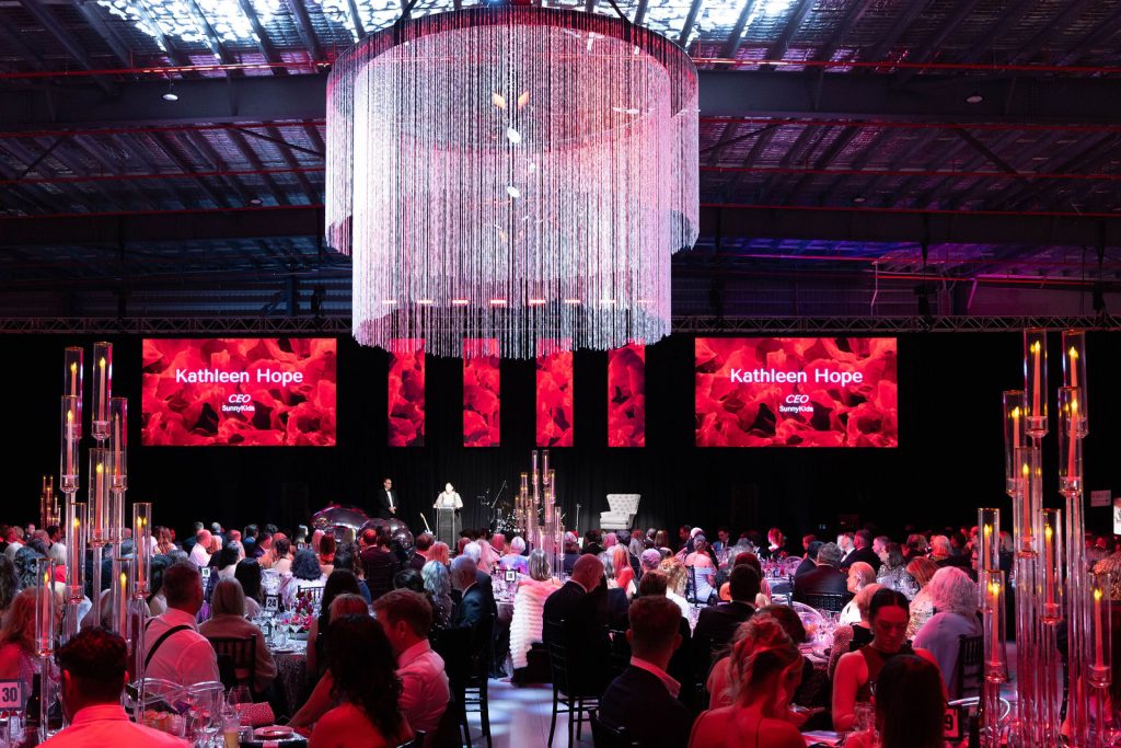 SunnyKids CEO Kathleen Hope speaks at the SunnyKids Mayoral Ball beneath a crystal chandelier, with guests seated at tables