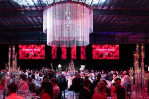 SunnyKids CEO Kathleen Hope speaks at the SunnyKids Mayoral Ball beneath a crystal chandelier, with guests seated at tables